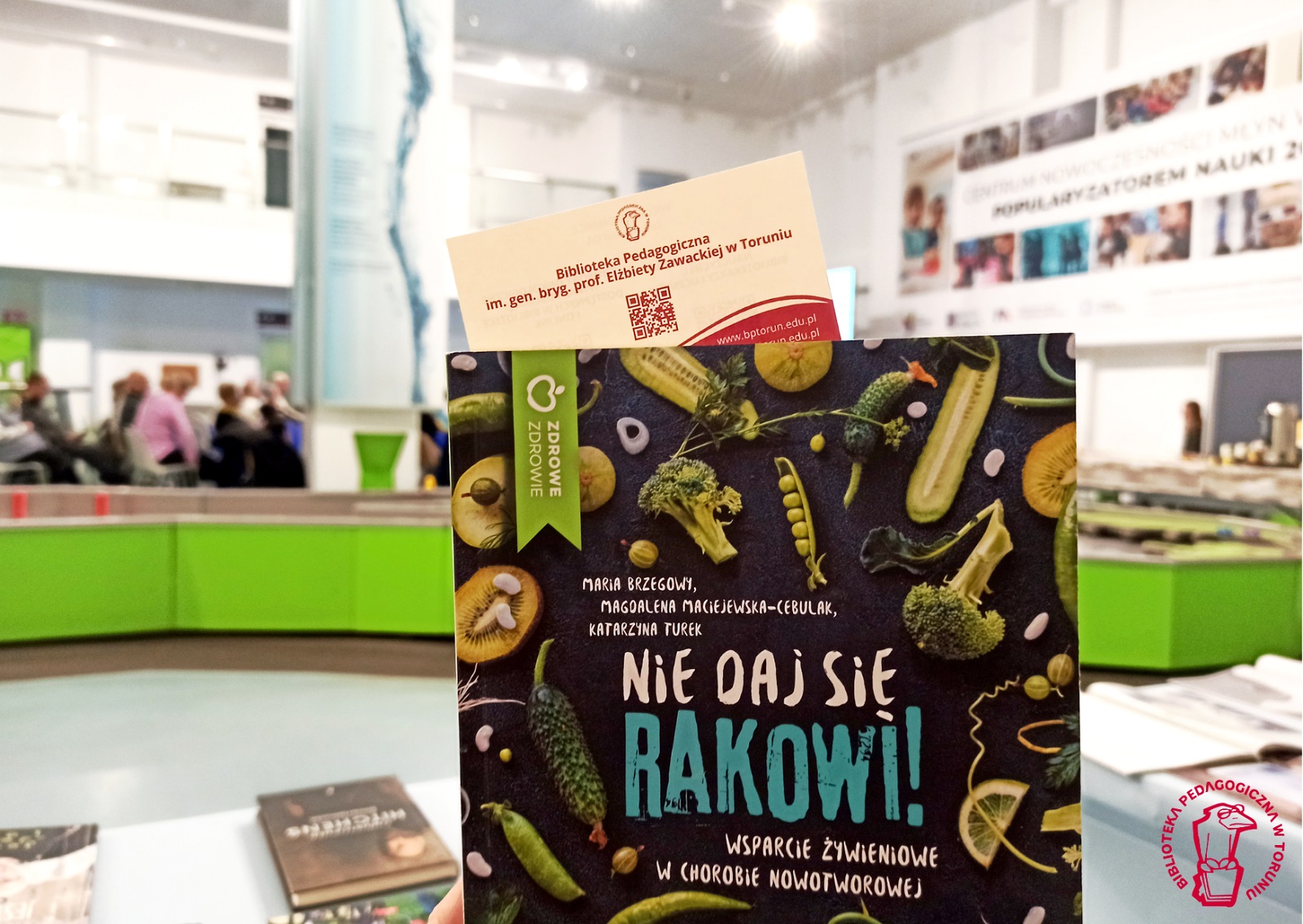 Okładka książki: Nie daj się rakowi! z motywami zielonych warzyw i owoców, na tle sali i publiczności w Młynie Wiedzy