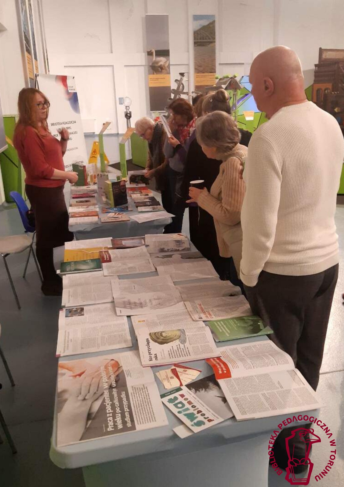 Kobiety i mężczyźni przy stoisku Biblioteki oglądają prezentowane na stołach książki i czasopisma