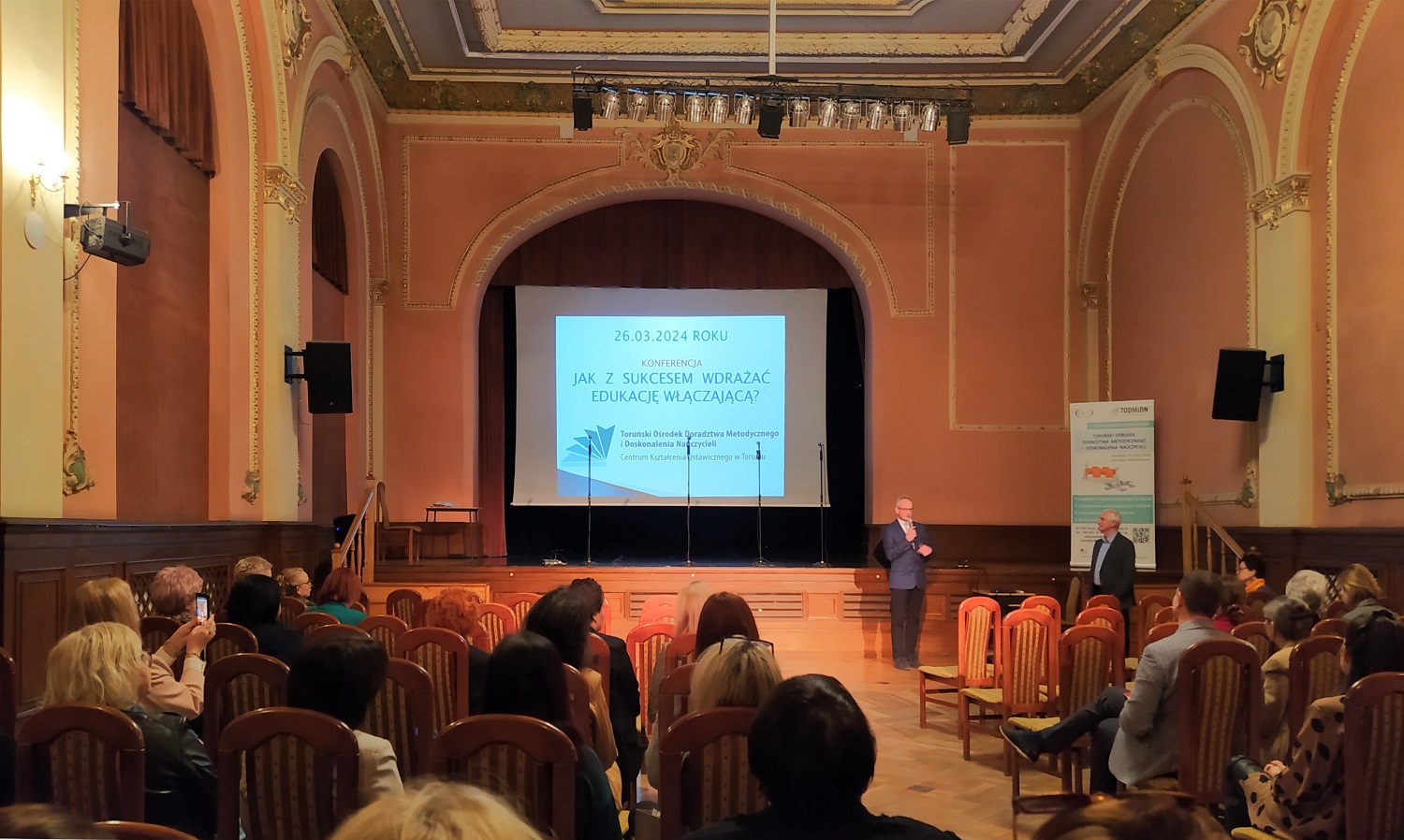 Duża, elegancka sala ze sceną. W centranym miejscu ekran z wyświetloną prezentacją. Liczne osoby siedzące na krzesłach, a blisko sceny stoi dwóch mężczyzn.