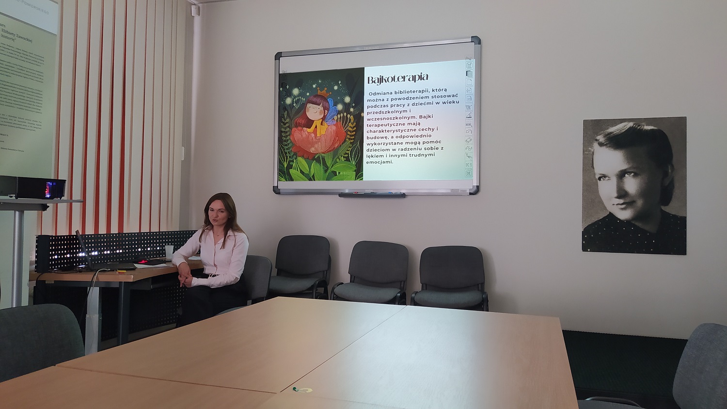 Długowłosa, ciemnowłosa kobieta siedzi przy komputerze. Nad nią na białej tablicy widoczny slajd prezentacji, zatytułowany: Bajkoterapia.