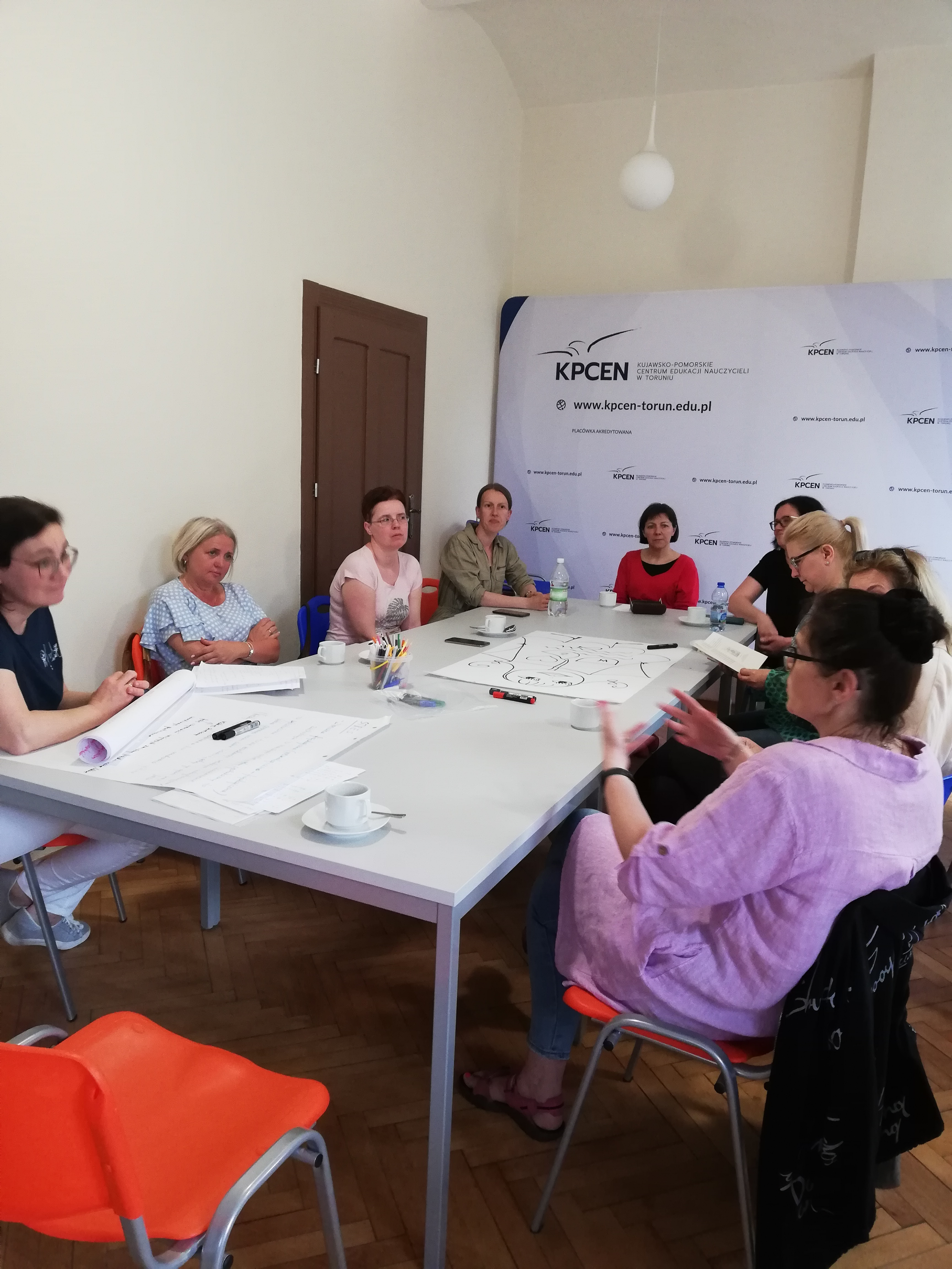 Uczestnicy spotkania siedzą przy stole w sali Kujawsko Pomorskiego Centrum Edukacji Nauczycieli w Toruniu. Za nimi stoi tablica informacyjna Kujawsko Pomorskiego Centrum Edukacji w Toruniu.