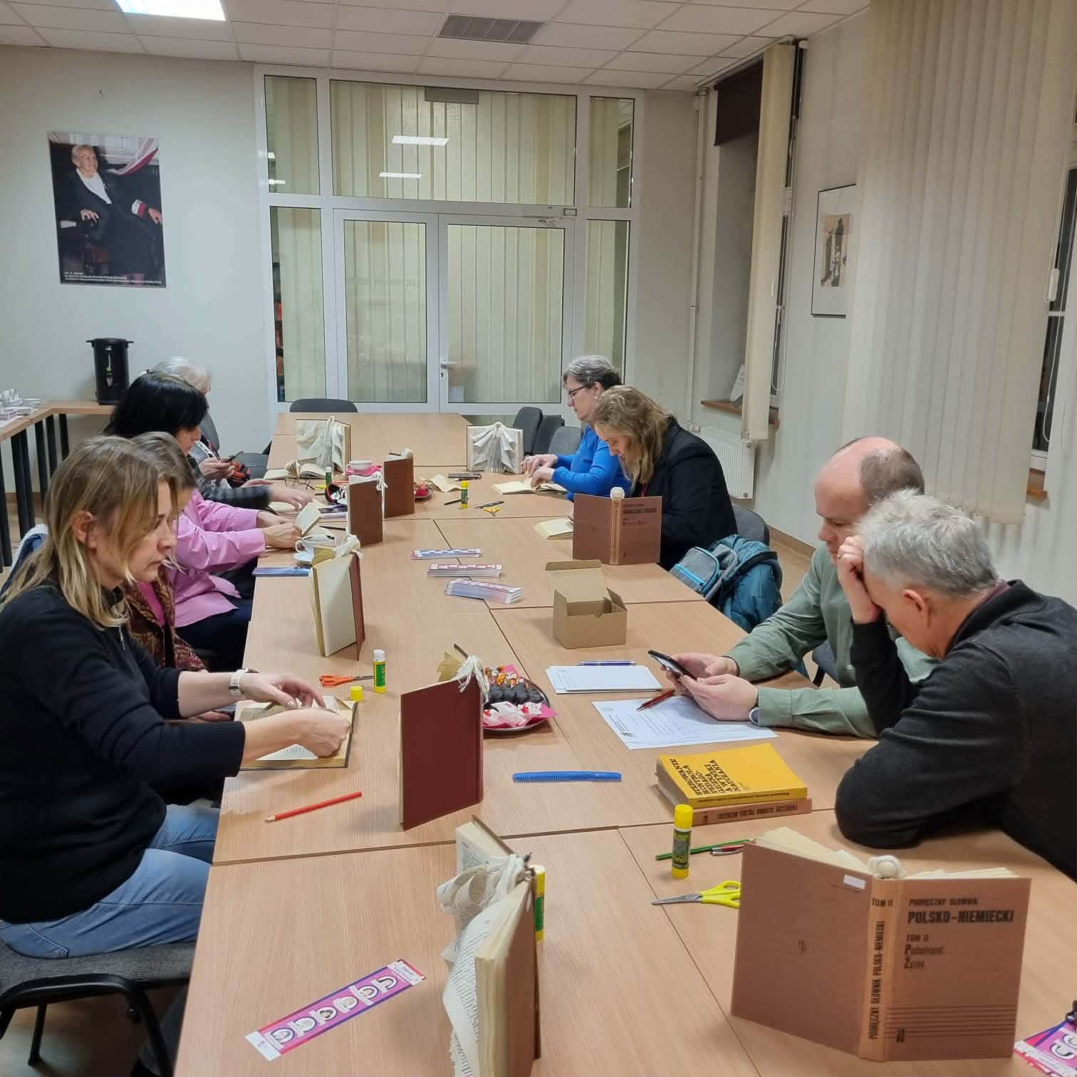 Grupa ludzi siedzi przy stołach w&nbsp;sali konferencyjnej Biblioteki Pedagogicznej w&nbsp;Toruniu. Przed nimi stoją stare książki, uczestnicy spotkania wykonują prace plastyczne.