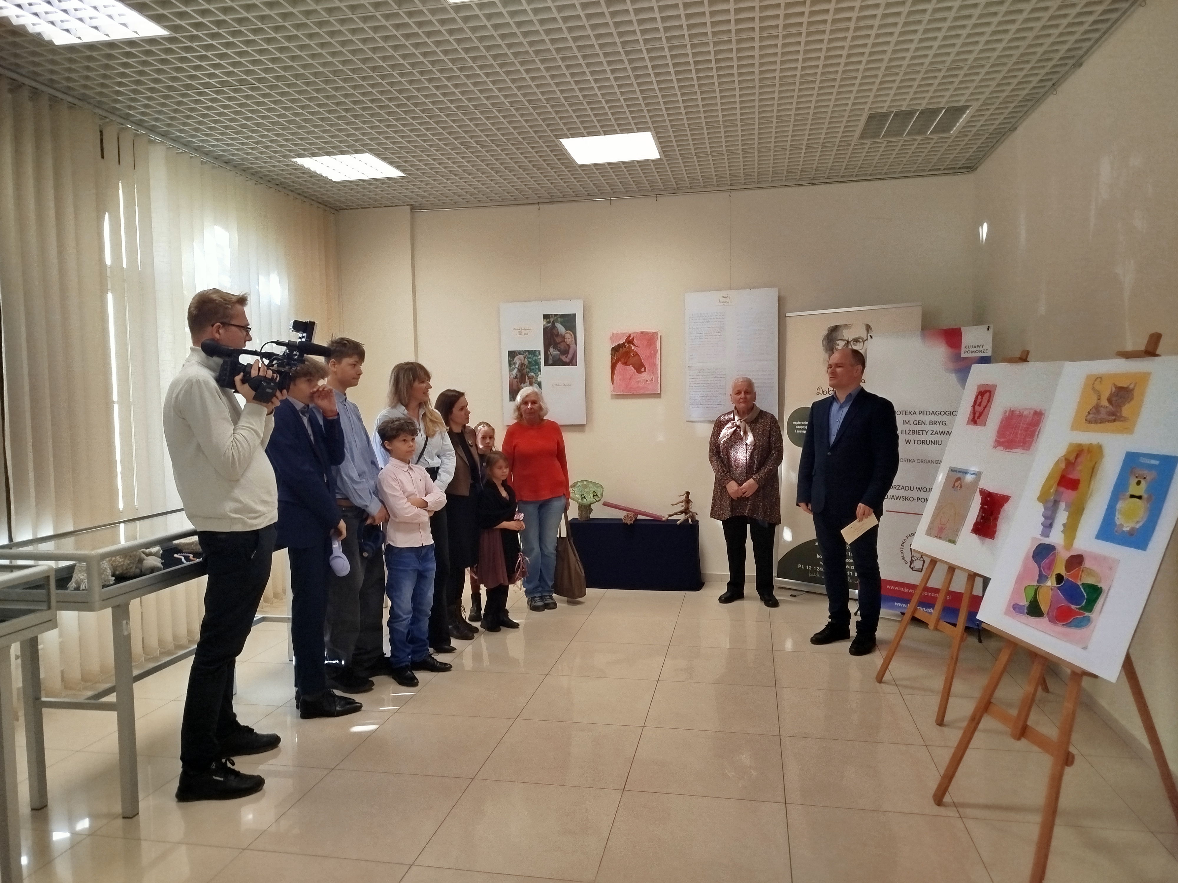 Grupa ludzi stoi w sali wystawowej Biblioteki Pedagogicznej w Toruniu.