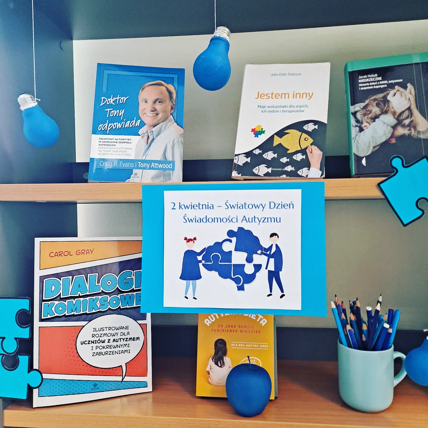 Zdjęcie przedstawia fragment regału z książkami na temat autyzmu. Widoczne elementy w kolorze niebieskim: żarówki, jabłko, kredki w niebieskim kubku.